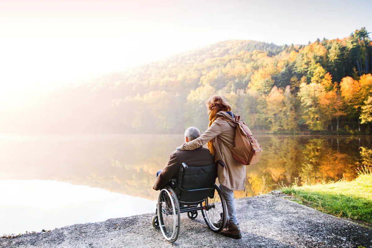 Couple de personnes aînées, dont une en fauteuil roulant, au bord d’un lac en automne